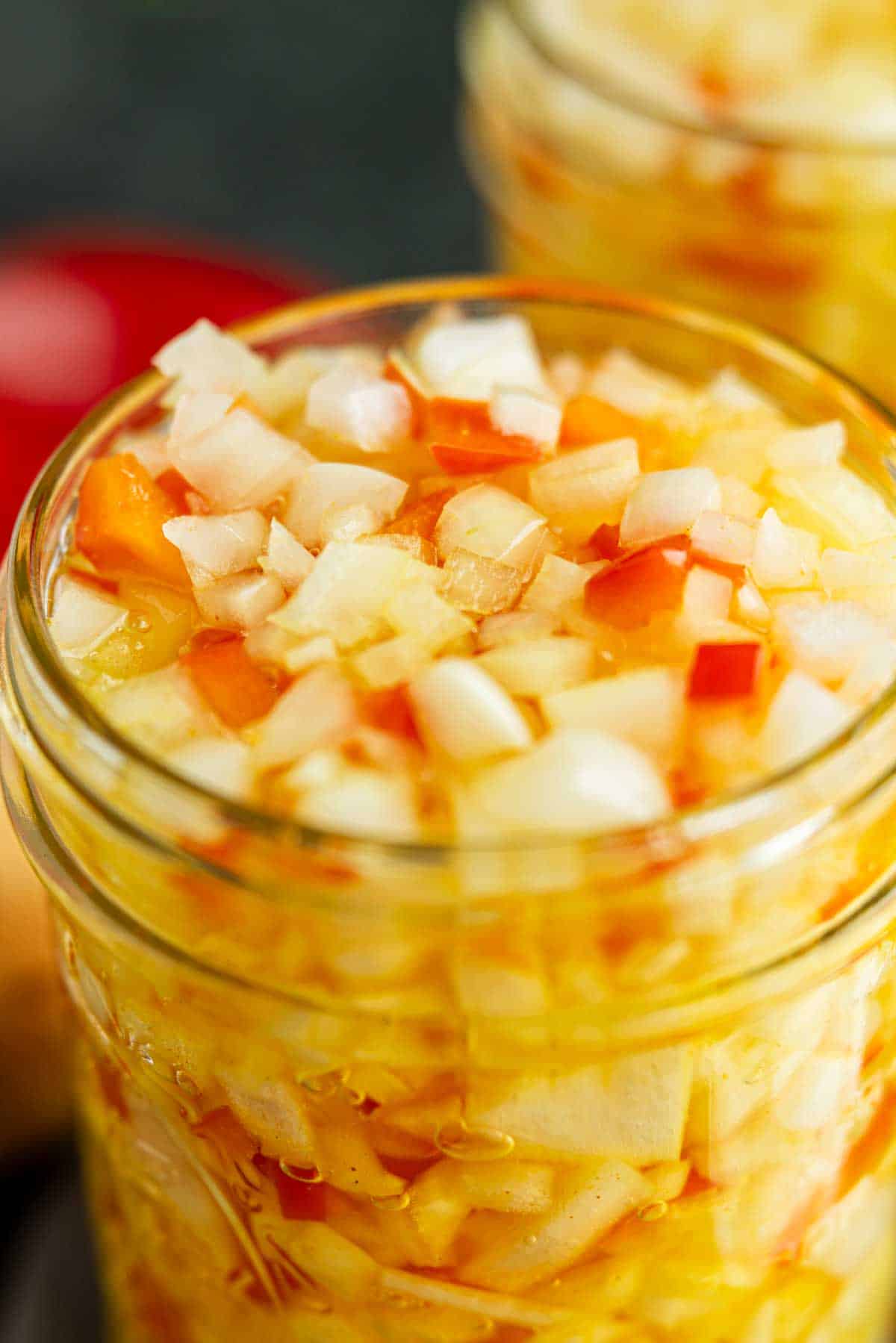 A close-up view of a glass jar filled with chopped onions and red peppers in a yellow liquid, likely a pickling brine.
