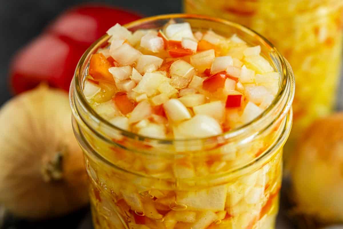 A close-up of a glass jar filled with chopped onions and red peppers in a liquid, with whole onions and a red bell pepper in the background.