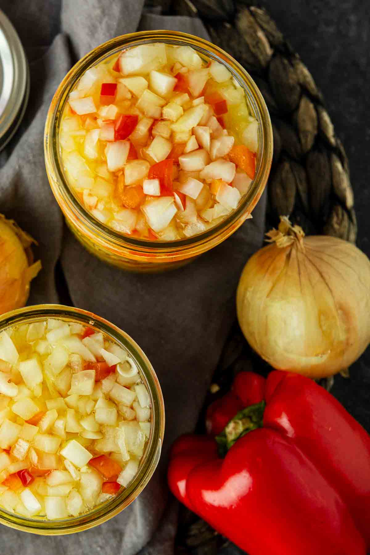Two jars of chopped pickled vegetables, including red peppers and onions, are placed on a cloth next to a whole onion and a red bell pepper.