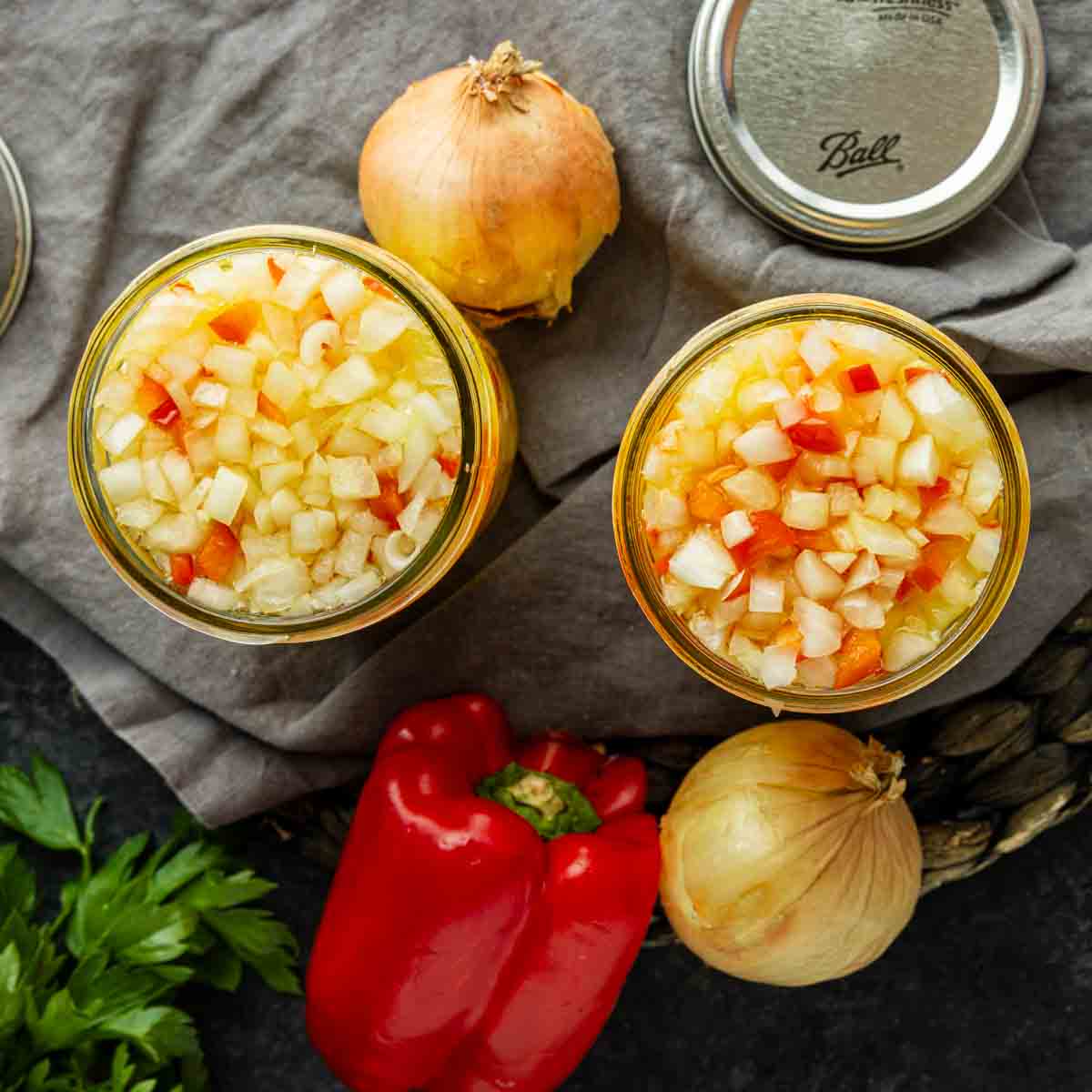 Two jars filled with chopped onions and red peppers on a cloth, surrounded by whole onions, a red bell pepper, parsley, and jar lids.