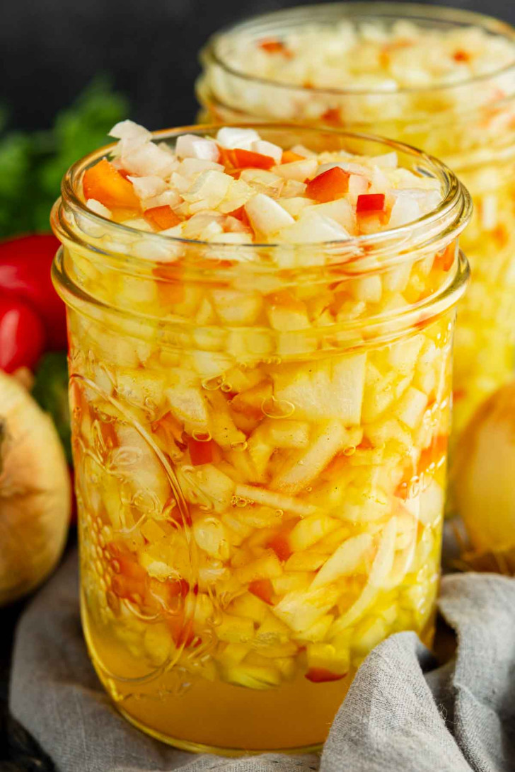 A close-up of two glass jars filled with diced onions and peppers in a yellowish liquid, with vegetables visible in the background.
