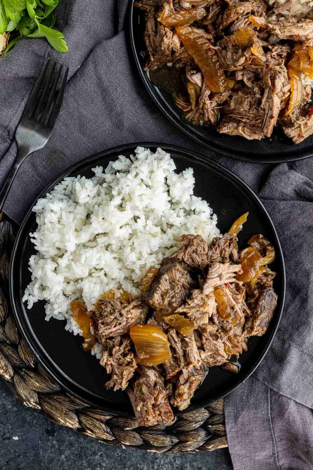 A black plate with white rice and shredded beef with onions, placed on a gray napkin with a fork and herbs nearby—this dish is inspired by an easy slow cooker dinner, featuring a flavorful chuck roast slow cooker recipe that’s both comforting and convenient.
