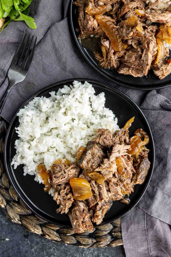 A black plate with white rice and shredded beef with onions, placed on a woven mat next to a fork and gray napkin.