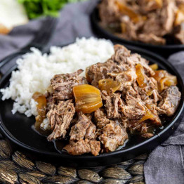 A black plate with shredded beef and cooked onions in sauce, served alongside white rice—this easy slow cooker dinner is a comforting meal perfect for any night of the week.