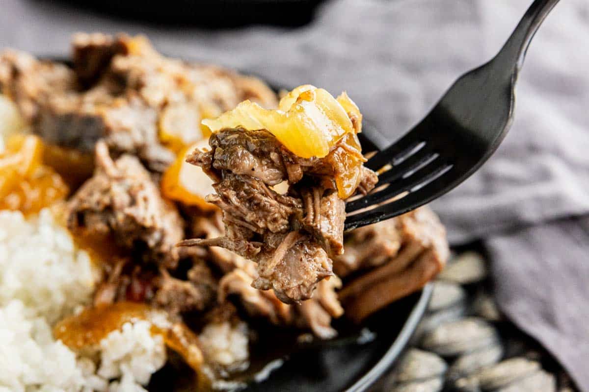 A black fork holds a bite of shredded beef with onions above a plate of beef and white rice, showcasing the rich flavors of this chuck roast slow cooker recipe.