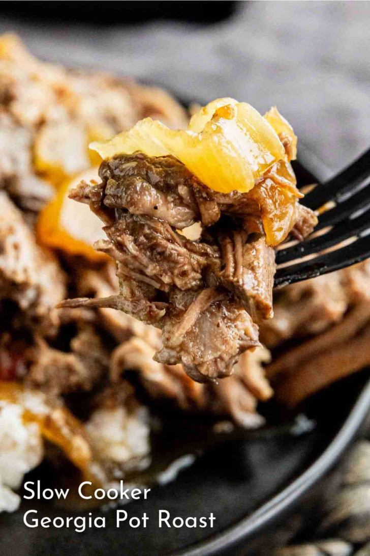 Close-up of a fork holding a bite of shredded pot roast with onions, with more pot roast on a plate in the background. Text reads “Slow Cooker Georgia Pot Roast.”.