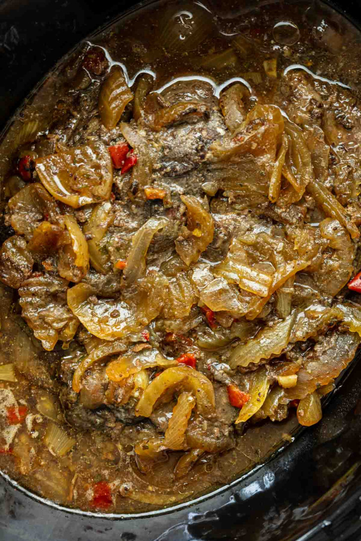 Close-up of cooked beef liver and onions with sliced peppers in a slow cooker, surrounded by a brown, savory sauce.