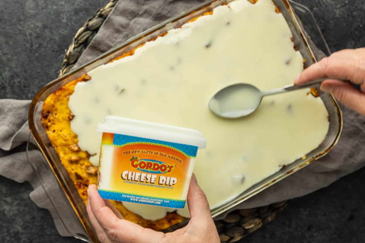 A hand spreads melted white cheese over a casserole in a glass dish with a spoon, while holding a container labeled “Gordo’s Cheese Dip”—a key step in preparing the viral TikTok chicken recipe known as swim queso chicken and rice.