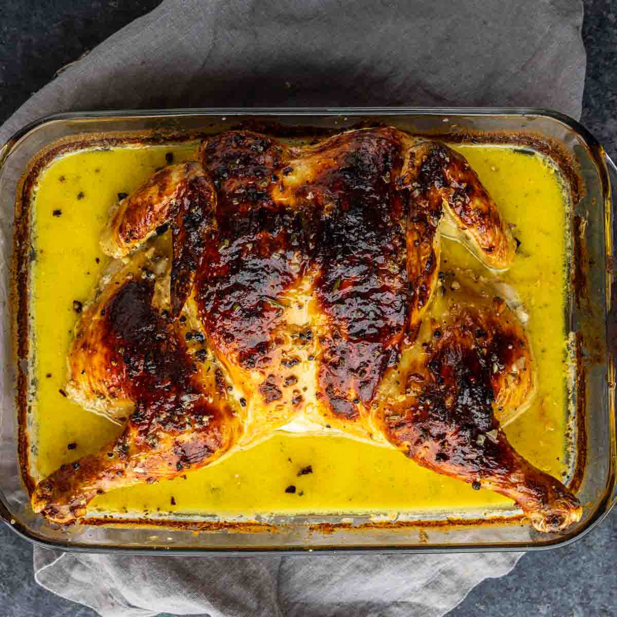 A whole roasted chicken, spatchcocked and marinated in a yellow sauce, sits in a glass baking dish on a dark cloth—a French-inspired chicken reminiscent of classic Chicken Au Poivre.