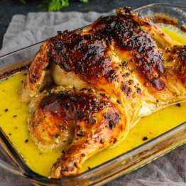 Roast spatchcock chicken with crispy skin and herbs sits in a glass baking dish with yellow pan juices.