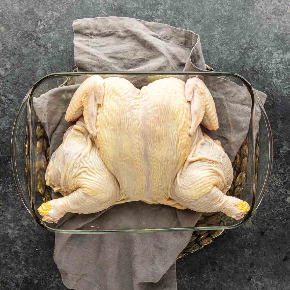 Raw spatchcocked chicken placed in a glass baking dish on a gray cloth and a wicker mat, ready for cooking preparation—perfect for creating a French-inspired chicken with peppercorn sauce, or classic Chicken Au Poivre.