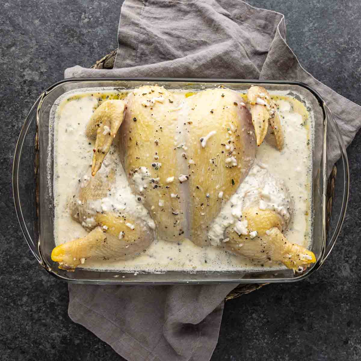 Raw spatchcocked chicken in a glass baking dish covered with a creamy white marinade, reminiscent of classic French-inspired chicken, placed on a gray cloth on a dark countertop.