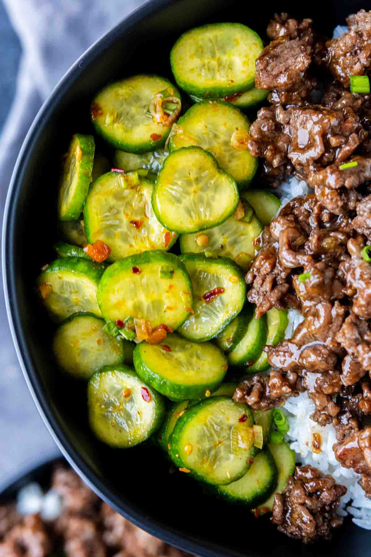 A bowl containing rice topped with seasoned ground beef and a spicy cucumber salad.