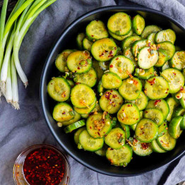 A black bowl filled with spicy cucumber salad topped with chili flakes and scallions, alongside green onions and a small dish of chili oil.