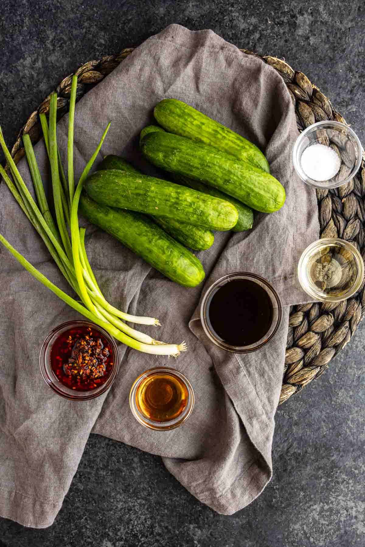 Cucumbers, scallions, and small bowls of chili oil, sesame oil, soy sauce, vinegar, and salt arranged on a linen cloth to make a spicy cucumber salad.