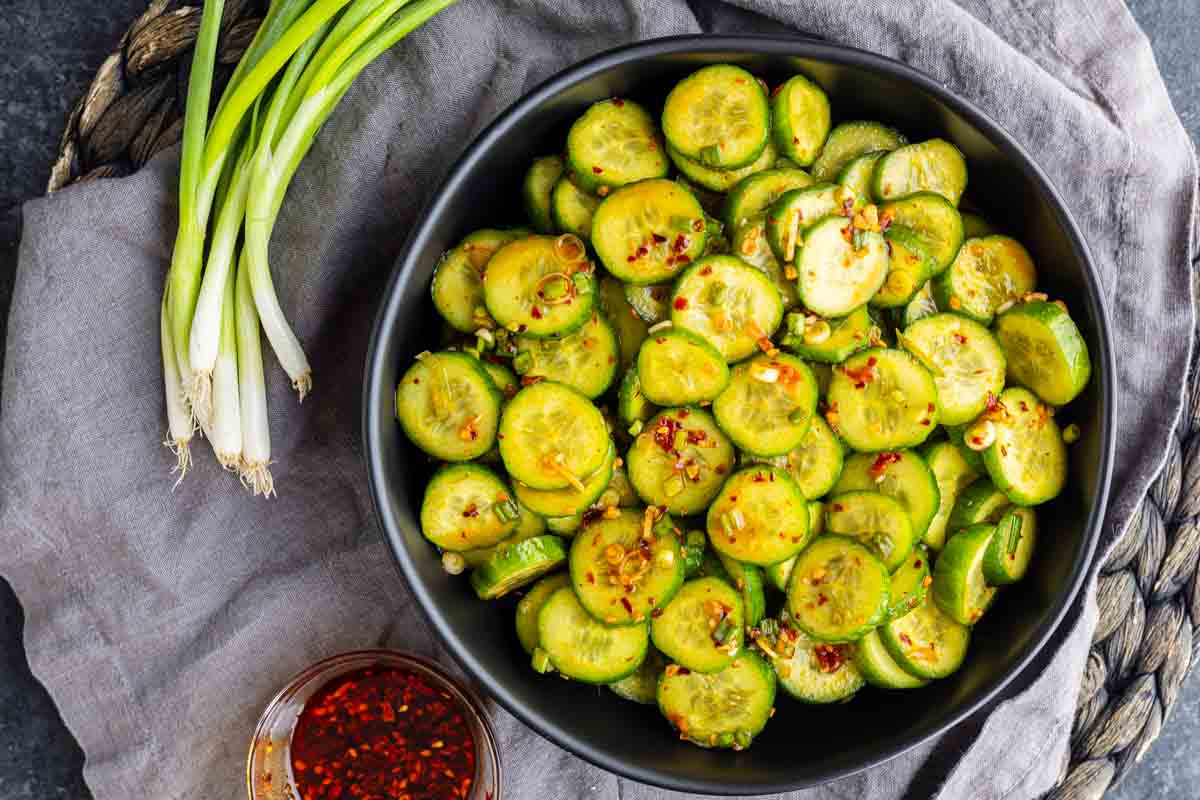 A black bowl filled with spicy cucumber salad topped with chili flakes, accompanied by a bunch of green onions and a small dish of chili oil.