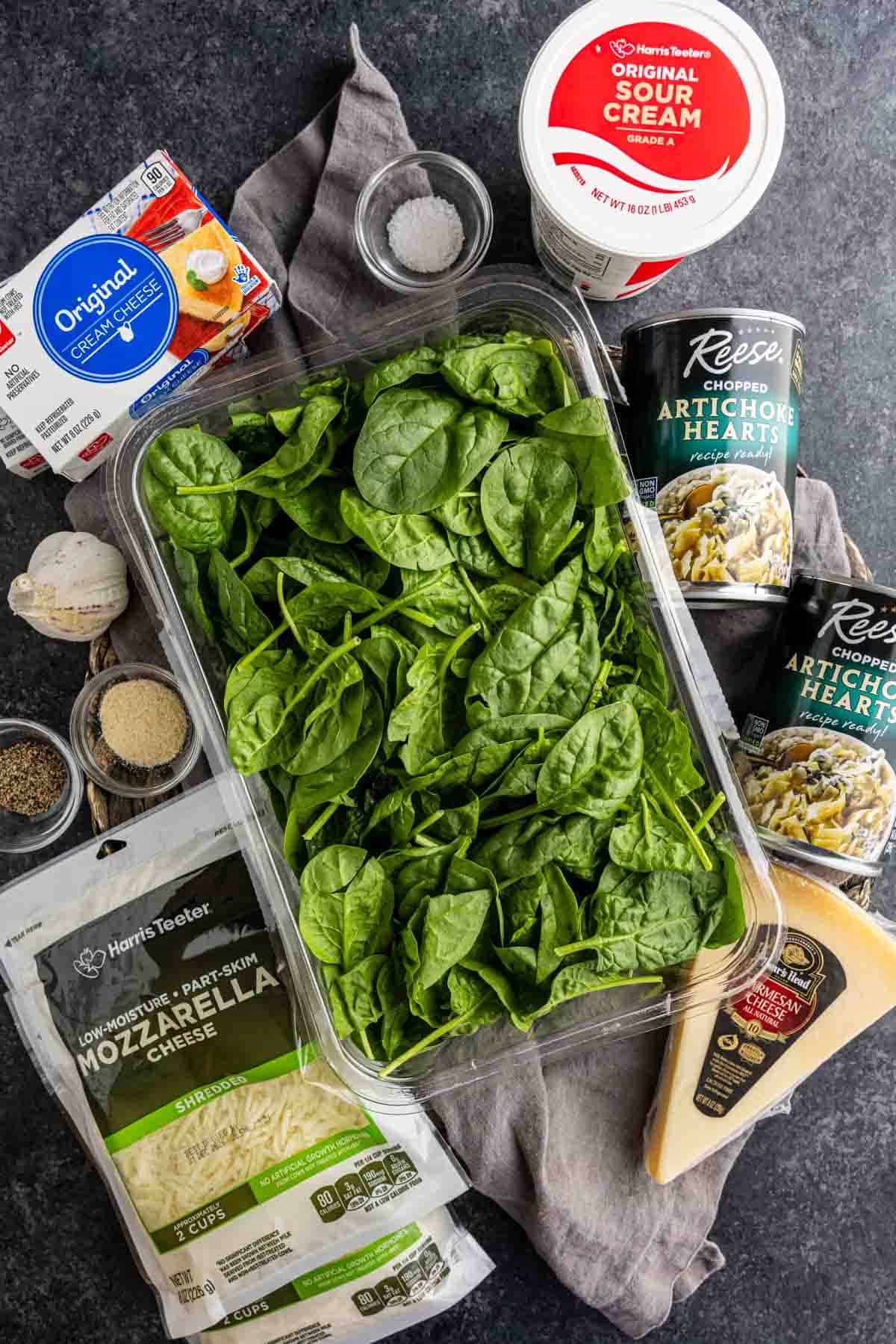 A tempting spread of ingredients for an easy crock pot spinach dip, featuring fresh spinach, sour cream, cream cheese, artichoke hearts, mozzarella, parmesan cheese, and seasonings—all beautifully arranged on a dark countertop for a delicious no mayonnaise option.