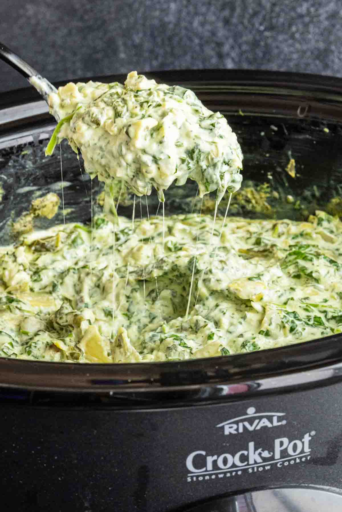 Spinach artichoke dip with fresh spinach is being lifted from a black slow cooker with a spoon, revealing gooey melted cheese and a creamy texture—this is a no mayonnaise slow cooker spinach artichoke dip everyone will love.
