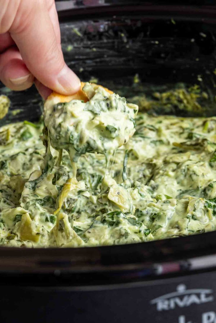 A hand dips a piece of bread into creamy slow cooker spinach artichoke dip with fresh spinach inside a slow cooker.