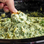 A hand dips a cracker into creamy spinach and artichoke dip with fresh spinach in a black dish, with cheese stretching from the dip—an irresistible no mayonnaise recipe that’s perfect for sharing.
