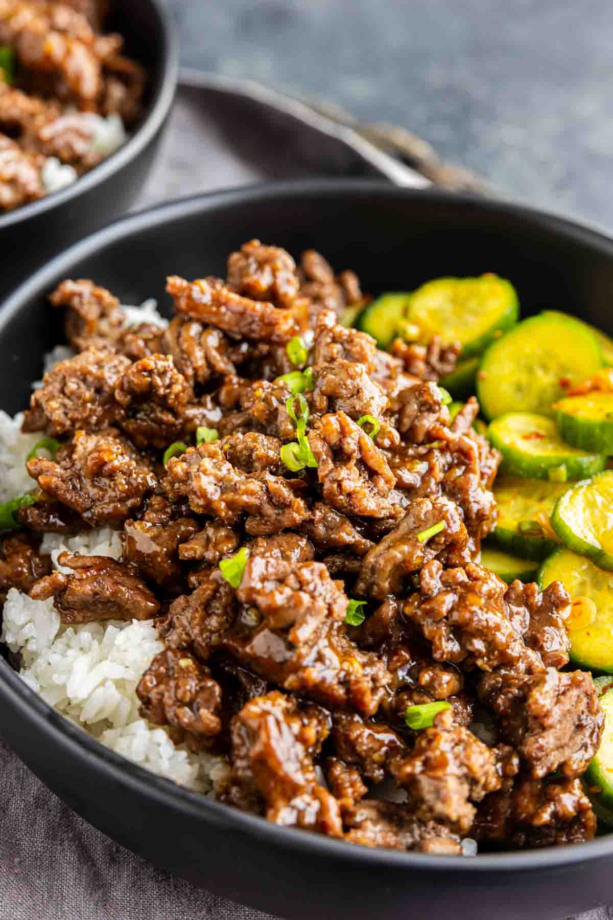 A black bowl filled with Korean beef bulgogi over white rice, served with a side of sliced cucumber salad for a delicious Korean ground beef bowl.