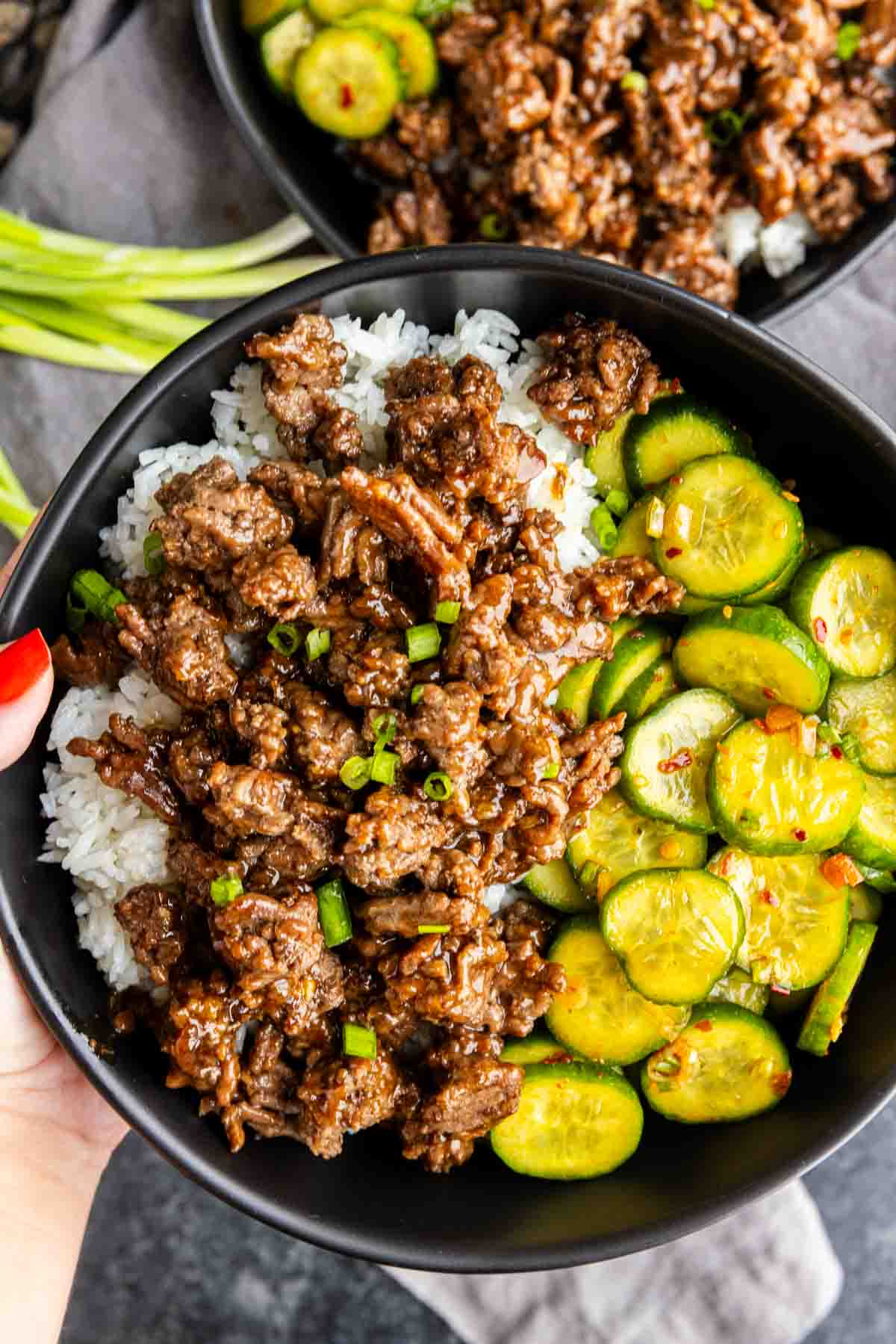 A bowl of Korean ground beef served over white rice with sliced cucumbers and a garnish of green onions.