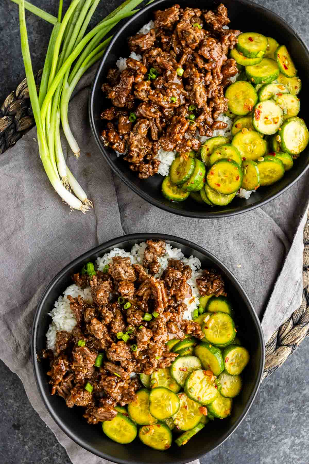 Two bowls of Korean ground beef and rice topped with sliced cucumbers and served with green onions on the side.