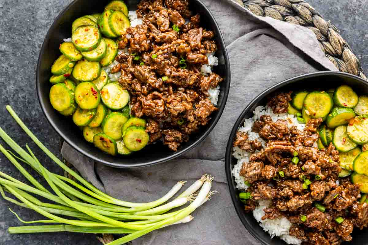 Two bowls of Korean ground beef and sliced cucumber over white rice, with a bunch of fresh green onions on the side.