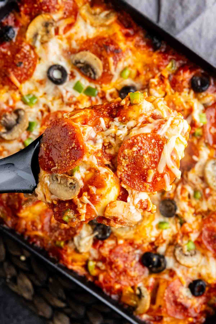 A close-up of a spatula lifting a serving of pizza casserole with melted cheese, pepperoni, mushrooms, olives, and green onions.