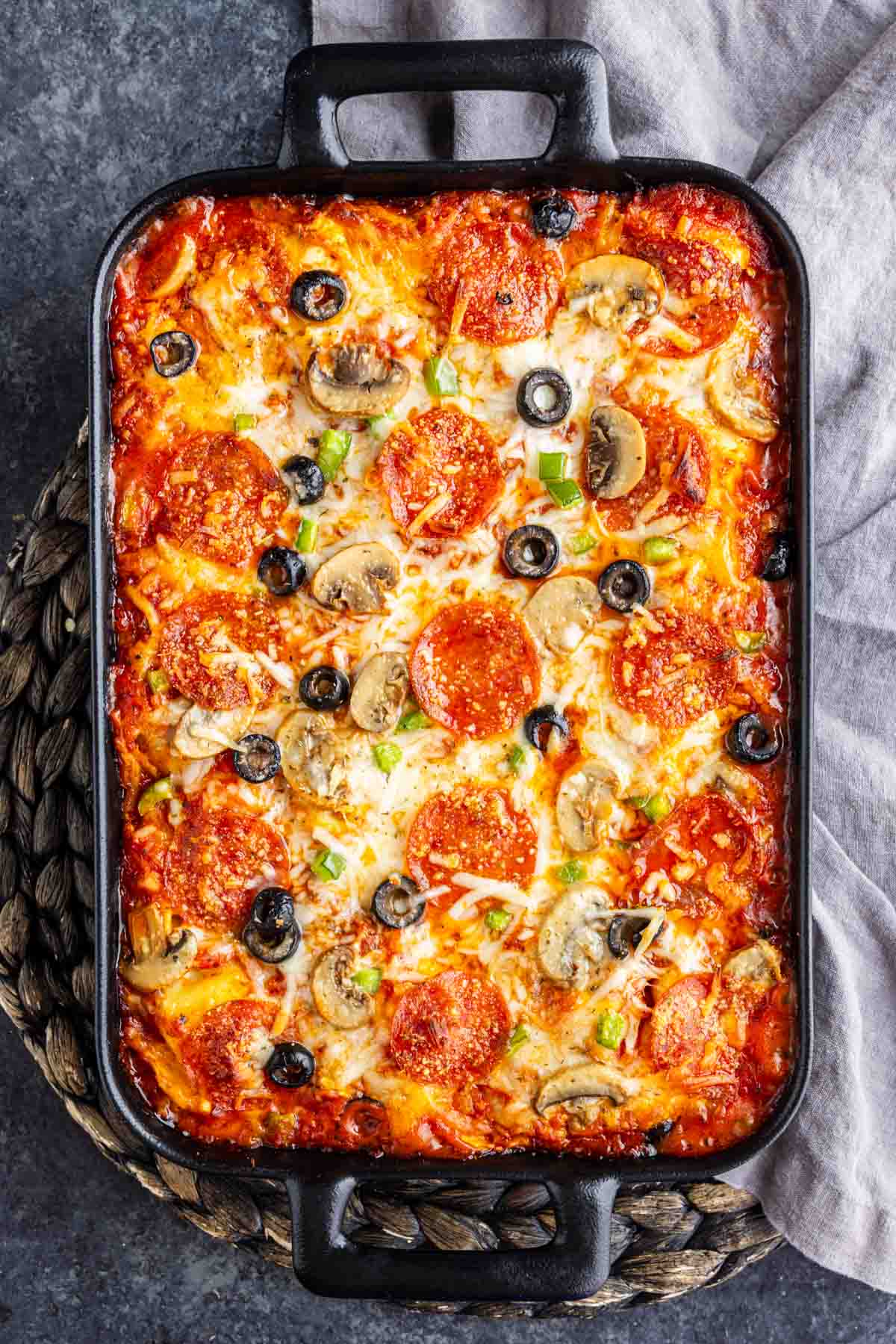 Rectangular baked pizza tortellini casserole topped with melted cheese, pepperoni slices, mushrooms, black olives, and green herbs, served in a black baking dish on a woven mat—an easy weeknight dinner the whole family will love.