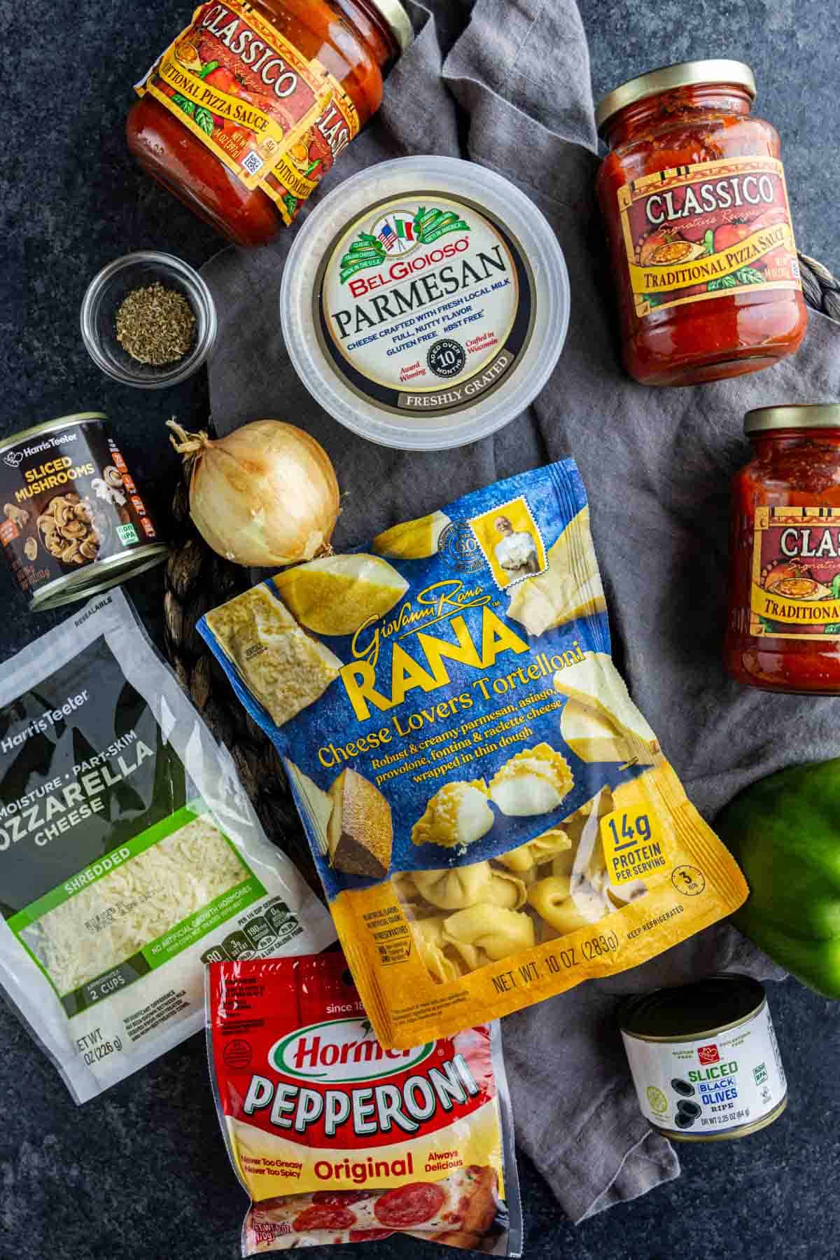 Assorted pizza ingredients on a dark surface, including baked pizza tortellini, sauces, parmesan, mozzarella, pepperoni, olives, onion, and green pepper.