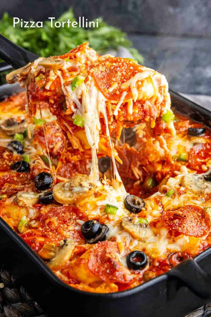 A pan of baked pizza tortellini topped with melted cheese, pepperoni, black olives, mushrooms, and green peppers, with a serving being lifted by a spatula.