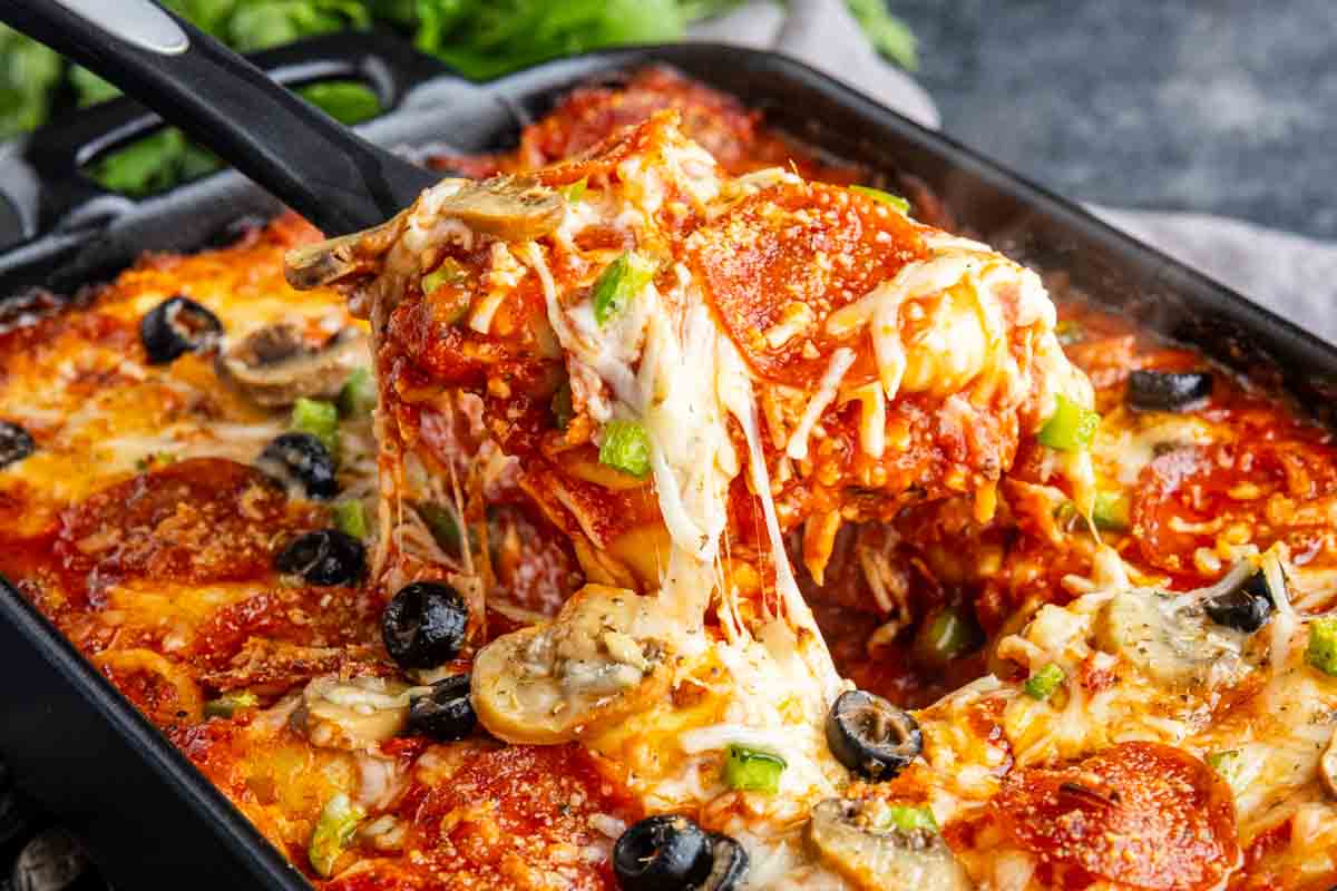 A spatula lifts a cheesy slice of pizza tortellini casserole from a pan, topped with pepperoni, mushrooms, olives, and green peppers.