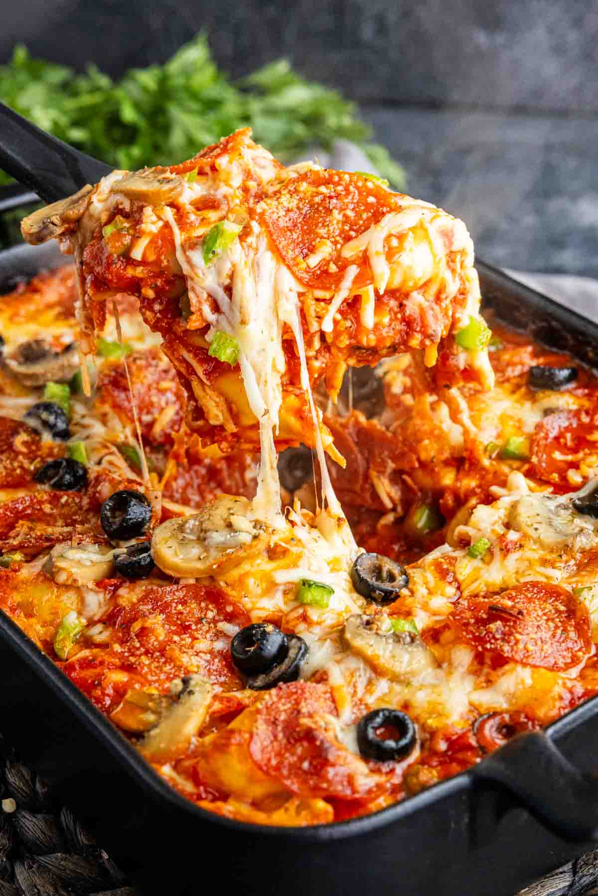 A cheesy baked casserole featuring pepperoni, olives, mushrooms, green peppers, and tomato sauce is being served with a spatula—making this baked pizza tortellini the perfect easy weeknight dinner.