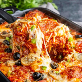 Close-up of a cheesy baked casserole with pepperoni, olives, mushrooms, green peppers, and tomato sauce being served from a baking dish—this irresistible pizza tortellini bake is perfect for an easy weeknight dinner.