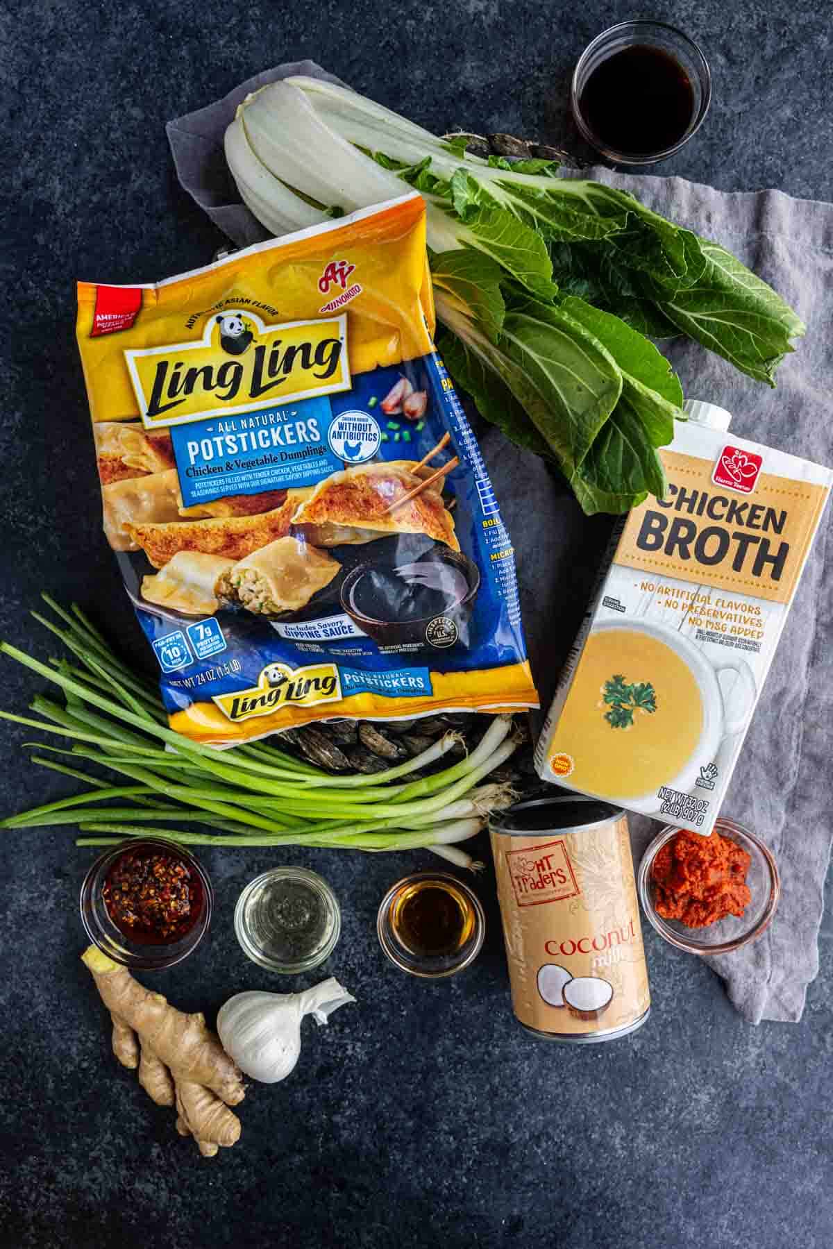 Assorted ingredients on a dark surface, including Ling Ling potstickers, chicken broth, bok choy, coconut milk, green onions, ginger, garlic, soy sauce, and red curry paste—everything you need to make a delicious coconut curry dumpling soup inspired by the viral TikTok dumpling soup trend.