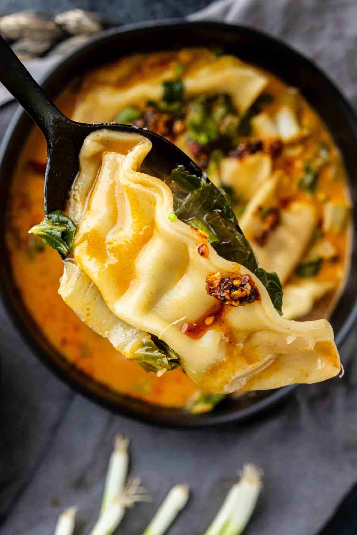 A spoon holds a dumpling above a bowl of soup with broth, greens, and chili oil, garnished with chopped vegetables—just like the viral dumpling soup trend taking over TikTok.