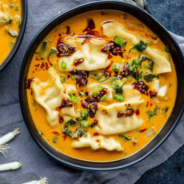 A bowl of creamy orange broth with dumplings, chopped green onions, and a drizzle of chili oil, placed on a gray cloth—a vibrant coconut curry dumpling soup that’s sure to become your next viral dumpling soup obsession.