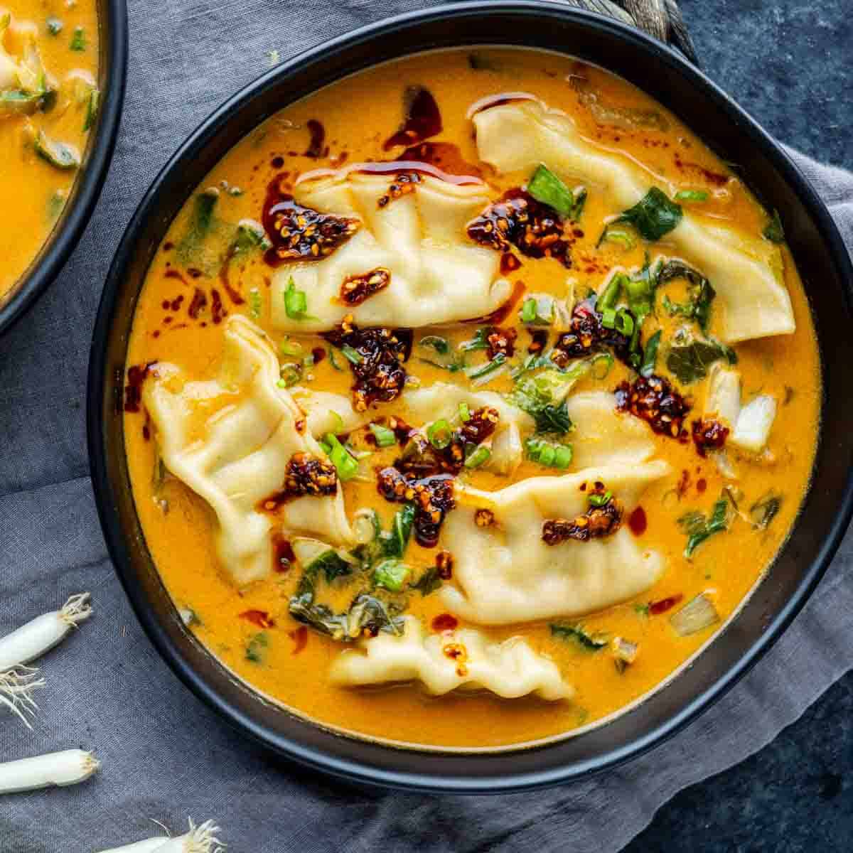 A black bowl filled with dumplings in a creamy orange broth, topped with chopped green onions and chili oil, on a gray cloth—this is the irresistible red curry dumpling soup that’s gone viral as the latest viral dumpling soup sensation.