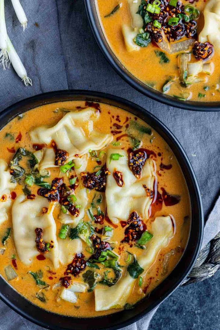 Two black bowls filled with dumplings in a creamy orange broth, garnished with chopped green onions and chili oil, placed on a dark cloth.