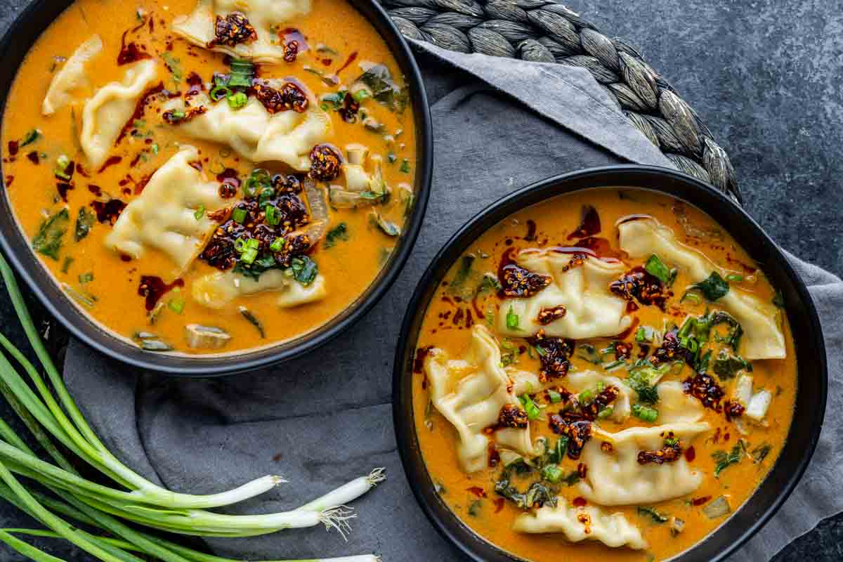 Two bowls of red curry dumpling soup with a creamy orange broth, garnished with chili oil, green onions, and greens, sit on a gray cloth next to scallions and a woven mat—just like the viral dumpling soup trending on TikTok.