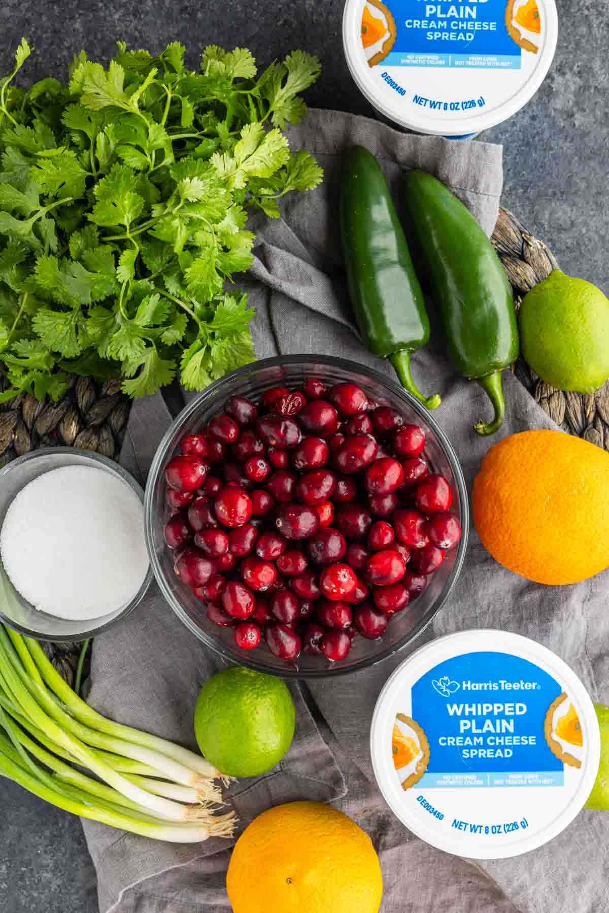 A glass bowl of ingredients for cranberry jalapeño dip, featuring fresh cranberries, cilantro, jalapeños, limes, oranges, green onions, sugar, and two containers of Harris Teeter plain cream cheese spread.