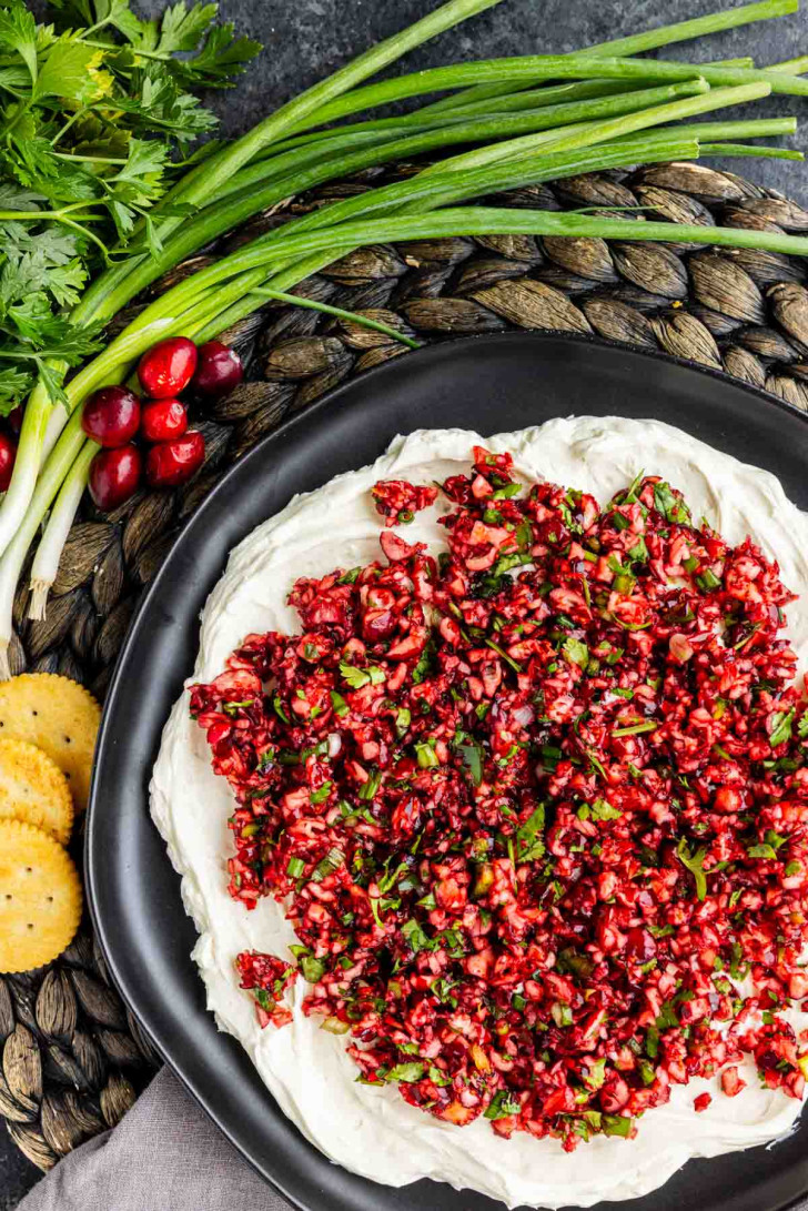A black plate topped with a creamy cranberry jalapeño dip covered in an herb salsa, served with round crackers, fresh cilantro, and green onions.