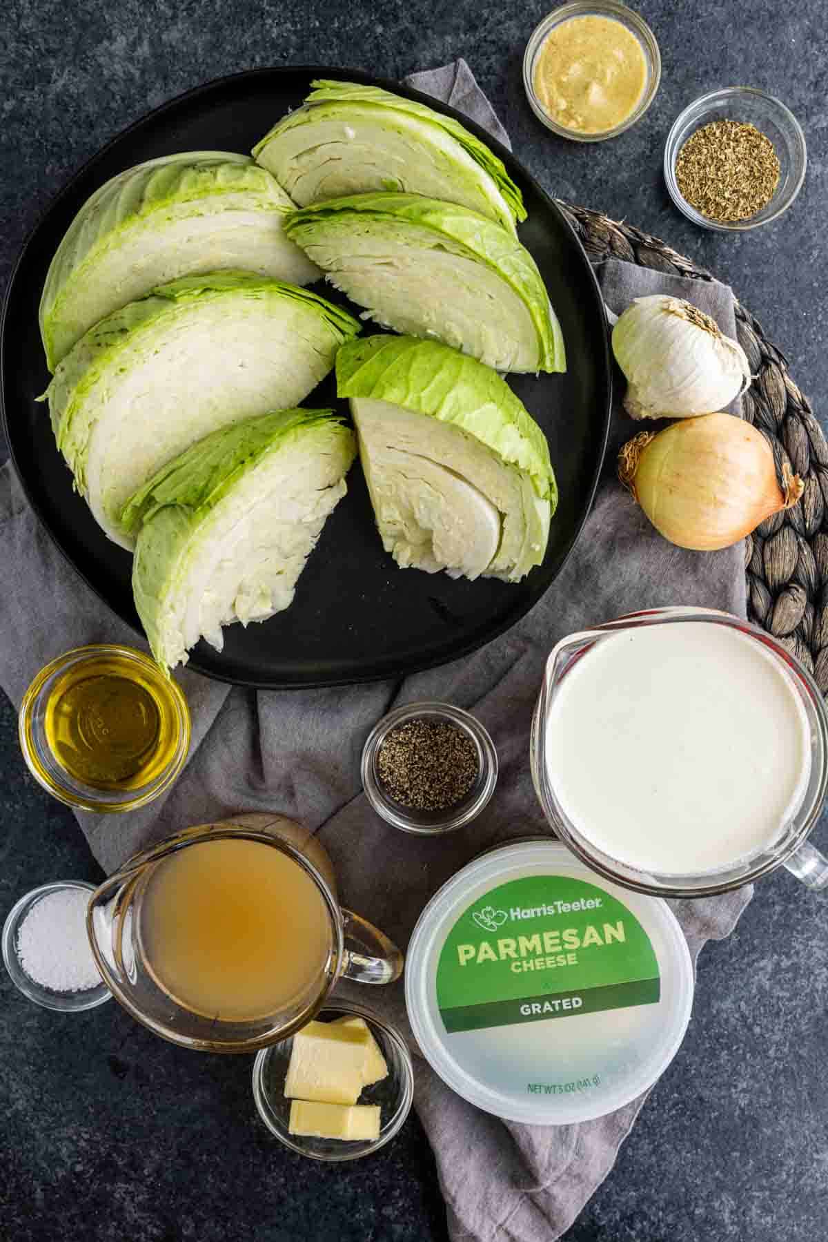 Six cabbage wedges for a creamy baked cabbage recipe arranged on a plate surrounded by garlic, onion, butter, oil, broth, heavy cream, parmesan cheese, and spices.