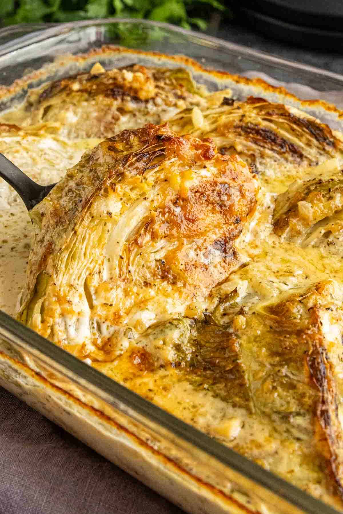 A spatula lifts a melting cabbage wedge from a creamy, baked casserole in a glass dish.