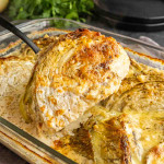 A serving spoon lifts a roasted cabbage wedge coated in a creamy, browned sauce from a glass baking dish.
