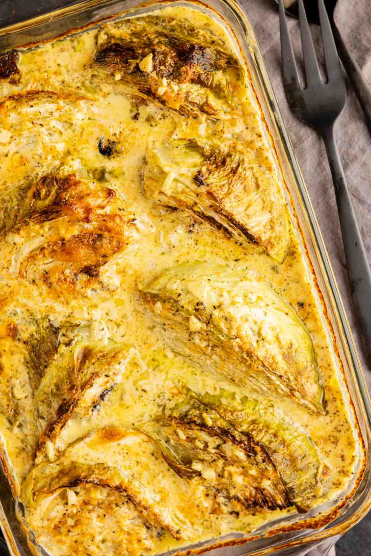 Baked cabbage wedges in a creamy sauce in a glass baking dish, served with a fork.
