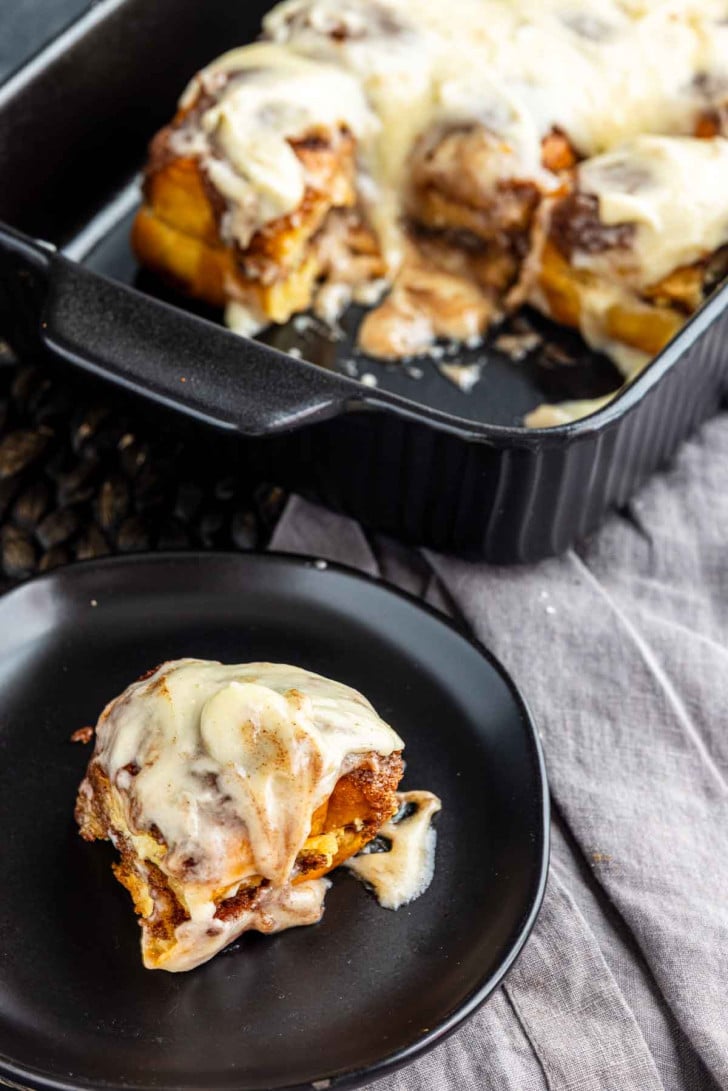 A frosted cinnamon roll on a black plate, with a baking dish of additional rolls in the background.