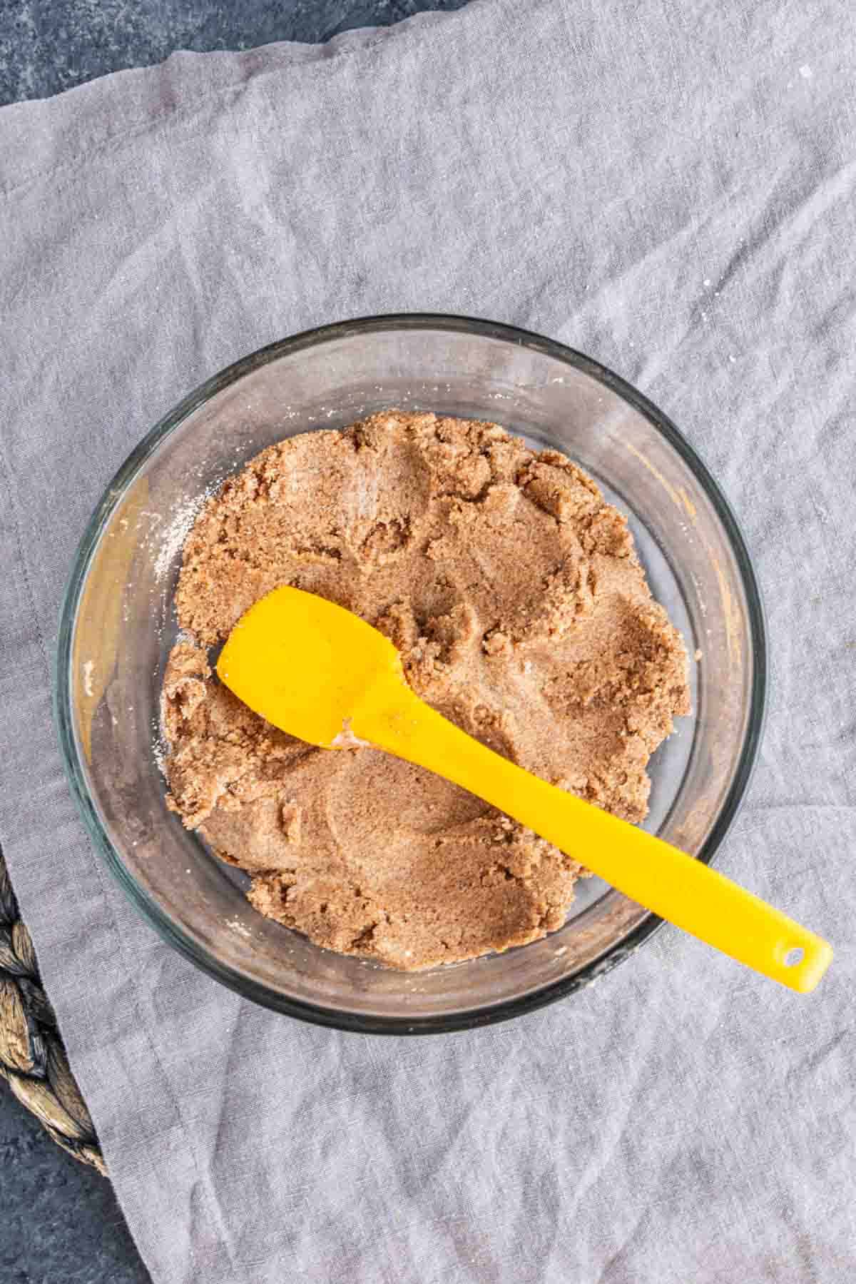 A yellow silicone spatula in a glass bowl filled with brown dough for King’s Hawaiian cinnamon rolls.