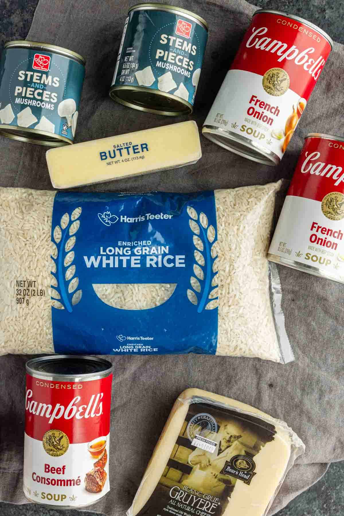 Ingredients for a French onion butter rice casserole: two cans of mushrooms, two cans of French onion soup, one can of beef consommé, a stick of butter, white rice, and a block of Gruyere cheese.